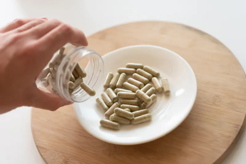 A hand pouring green kratom capsules from a clear plastic container onto a small white plate.