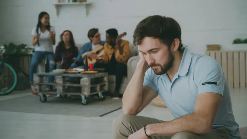 A young man sits in the foreground looking downward with an isolated and contemplative expression, while a group of friends happily socialize, play guitar, and drink from bottles in the blurred background. The image represents the social pressure problem and the subtle discomfort of being the only sober person at a gathering.