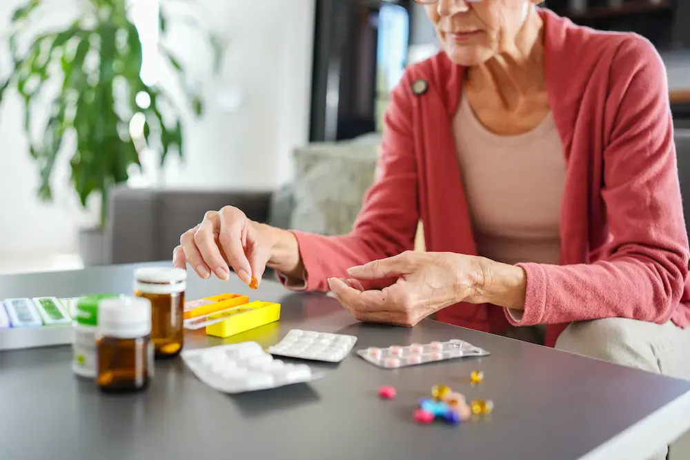An elderly person wearing a red cardigan sits at a table, carefully sorting a large collection of prescription medication, blister packs, and a pill organizer. The image illustrates the complex management of daily medications that can sometimes lead to accidental misuse or "medication fog" in seniors.