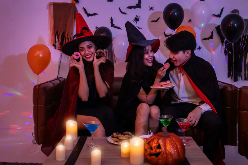 Three young adults in Halloween costumes enjoying donuts and colorful mocktails at a sober Halloween party.