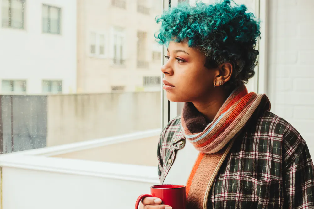 young-woman-with-blue-hair-looking-outside-at-winter-weather-feeling-sad