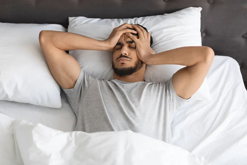 A young man lying in bed in a white shirt, holding his head with both hands with his eyes closed, appearing to suffer from a headache, stress, or withdrawal symptoms.