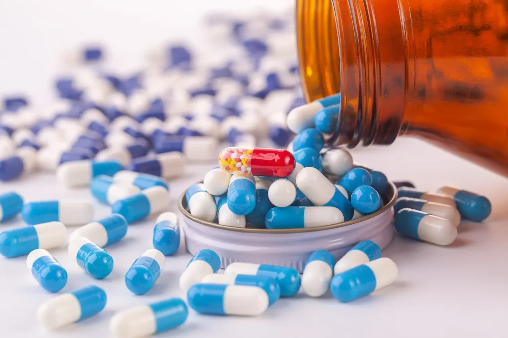 An amber medicine bottle spilled over on a white surface, scattering numerous blue and white capsules. Resting in the bottle's upturned cap among the blue pills is a single, distinct red and clear capsule filled with tiny colorful beads.