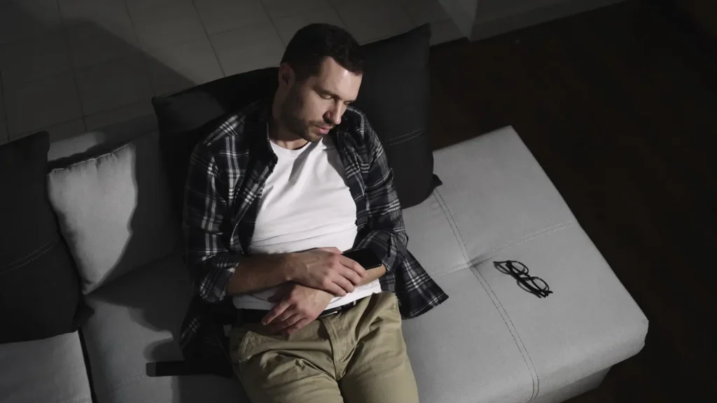 A man wearing a plaid shirt and khaki pants asleep on a gray sofa in a dark room, holding a smartphone with his glasses resting beside him.