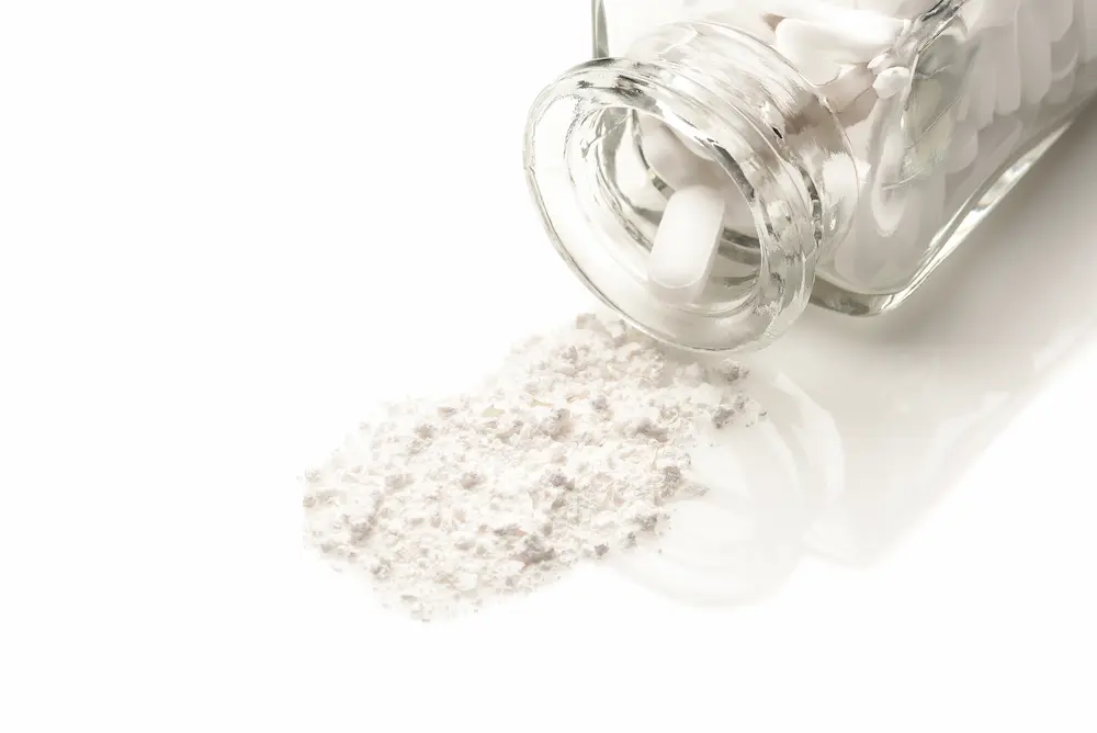 A clear glass bottle tipped on its side with white pills inside and white powder spilling out, illustrating the hidden danger of fentanyl-laced counterfeit medications.