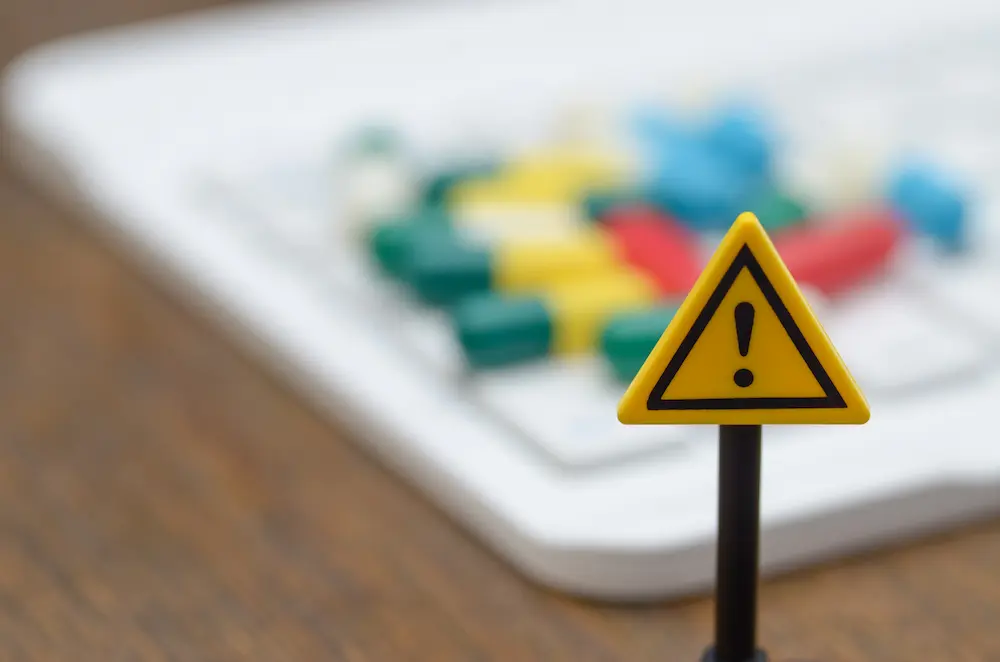 A small yellow warning triangle with a black exclamation point stands in focus in the foreground, with brightly colored pills blurred in the background, symbolizing the hidden dangers of counterfeit medication.