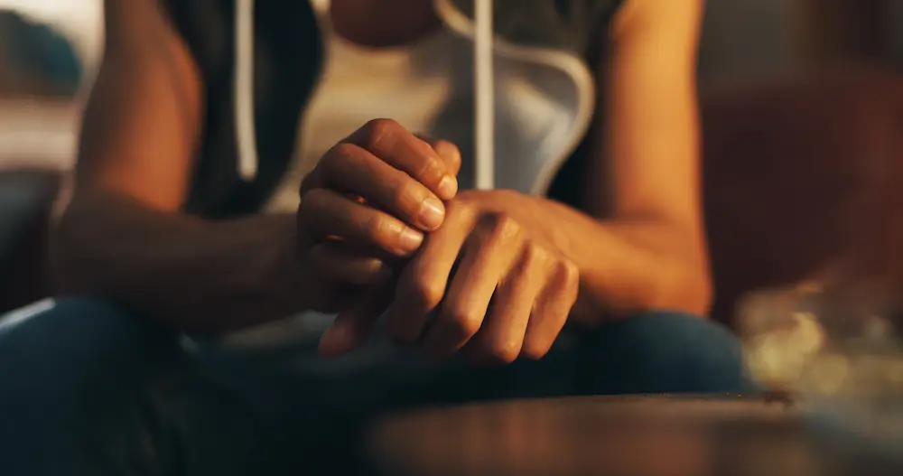 A close-up of a person sitting down, nervously scratching and rubbing their hands together, illustrating the severe physical restlessness and anxiety common during drug withdrawal.