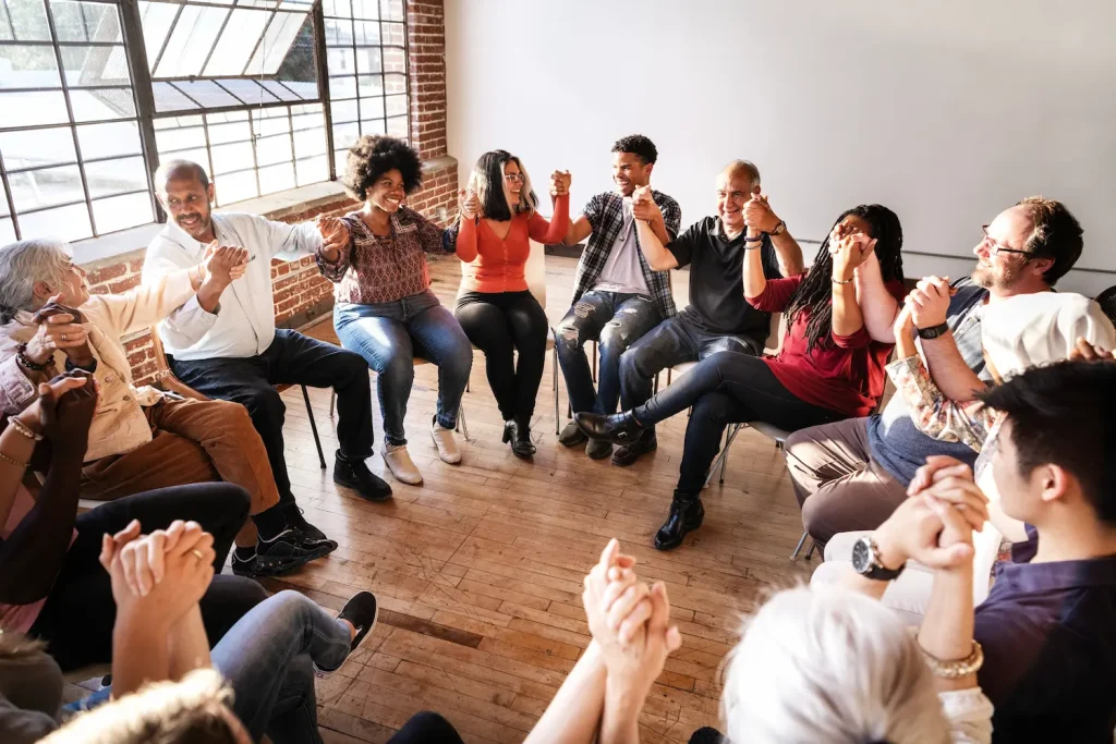 A diverse group of people sitting in a circle holding hands and smiling during a support group meeting.