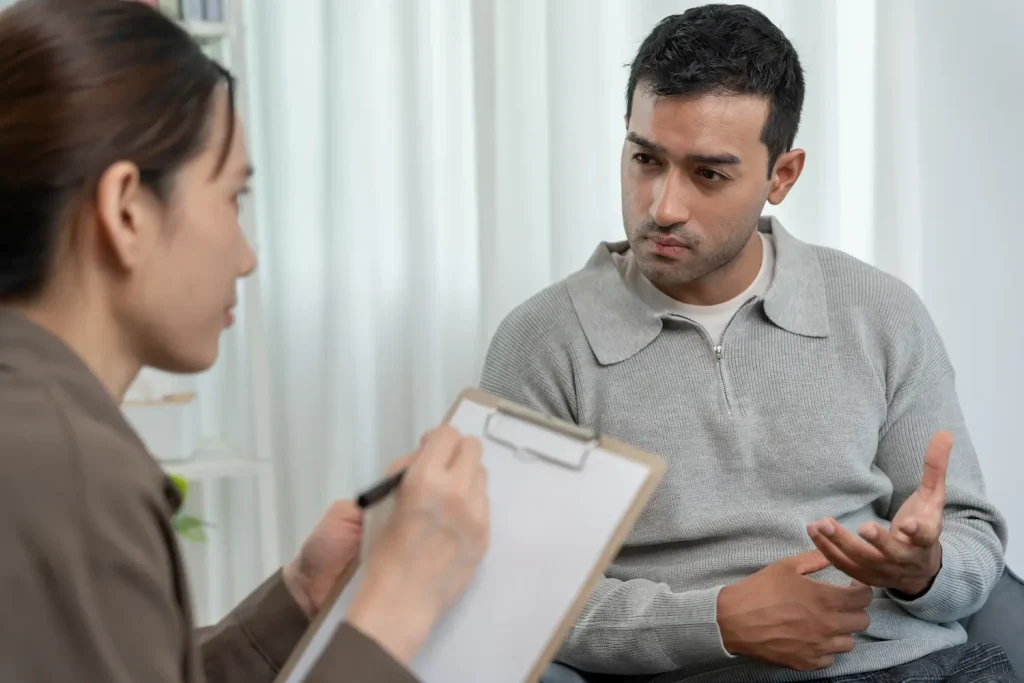 A male patient talking and gesturing while a female therapist takes notes on a clipboard during a counseling session.