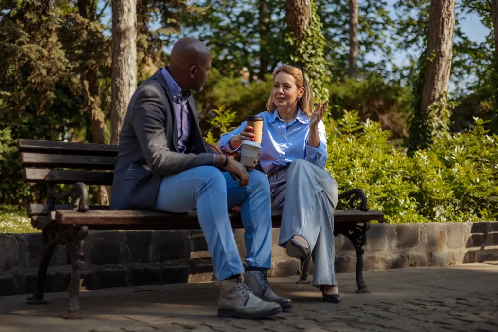 A man and a woman sit together on a sunny park bench, holding coffee cups and engaging in an earnest conversation. The image represents the importance of staying connected to a support network, such as checking in with a sponsor or a sober peer, during the high-risk summer months.