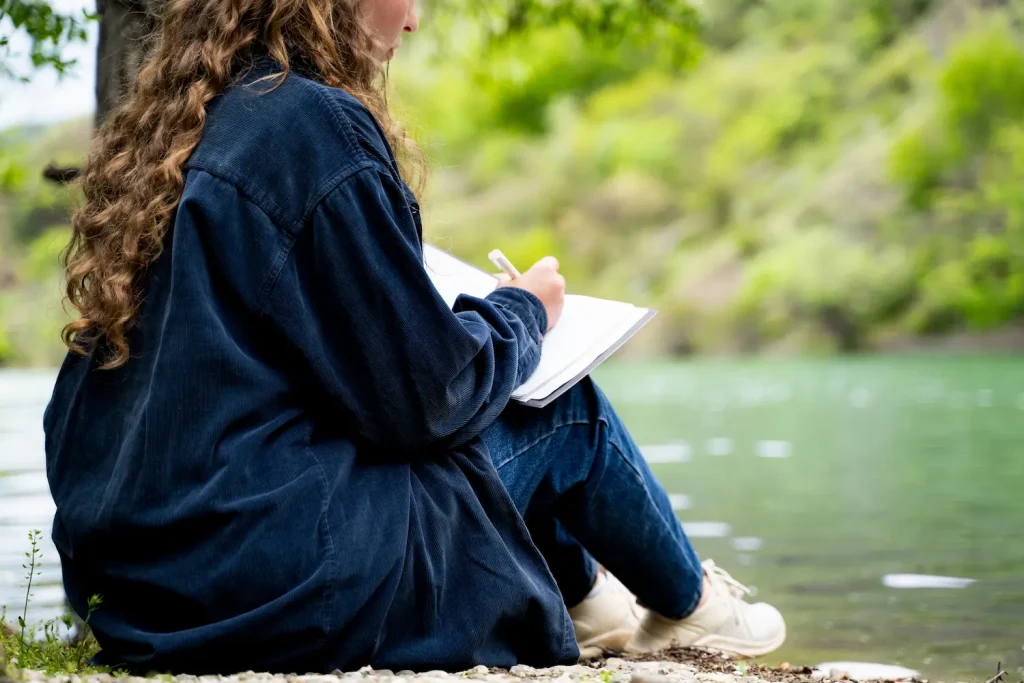 A woman with long curly hair sits peacefully on the bank of a calm river, writing in a notebook. The image represents the importance of deliberate planning, managing unstructured time, and finding quiet, relaxing sober activities during the summer.