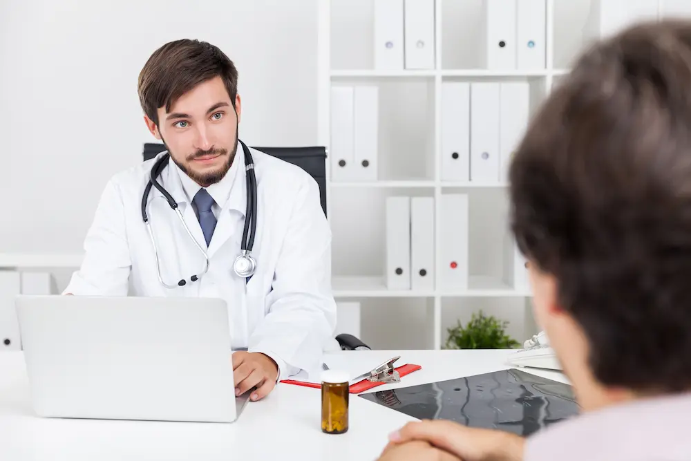 A young male doctor wearing a white coat and stethoscope sits at a bright office desk, looking attentively at a patient seated across from him whose back is to the camera. On the desk between them rests an open laptop, medical imaging films, a red clipboard, and a brown prescription pill bottle.