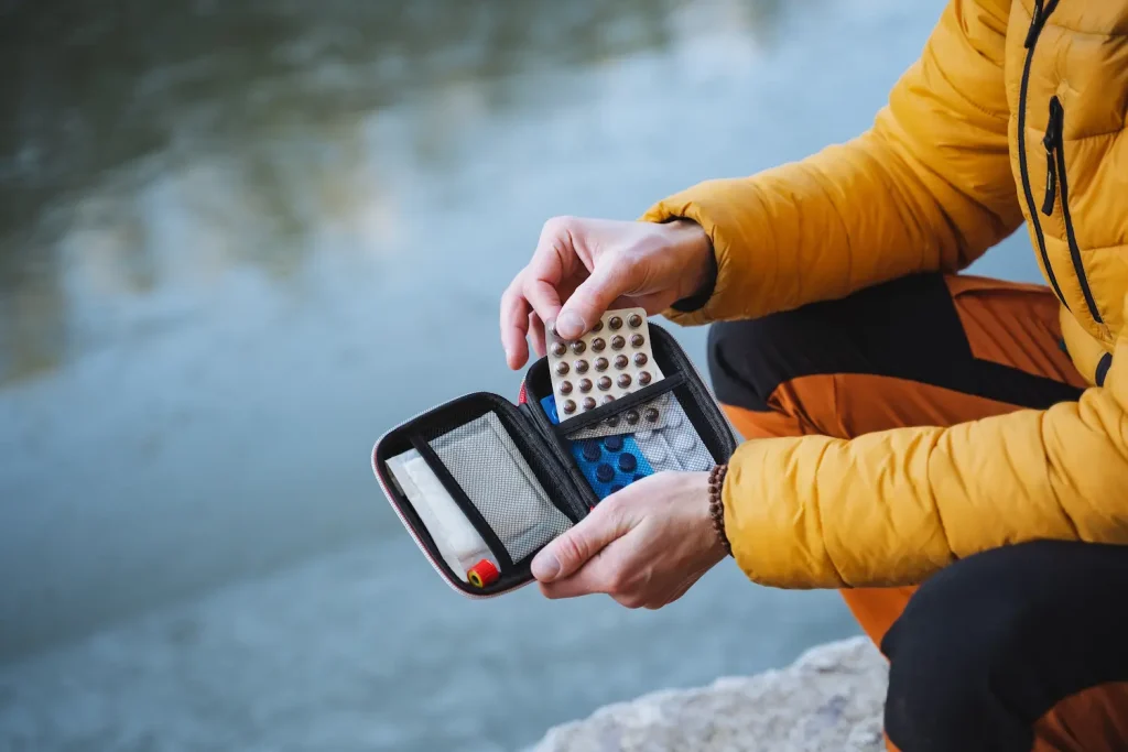 A person wearing a yellow jacket sits near a body of water, holding an open, small first-aid pouch and pulling out a blister pack of pills.