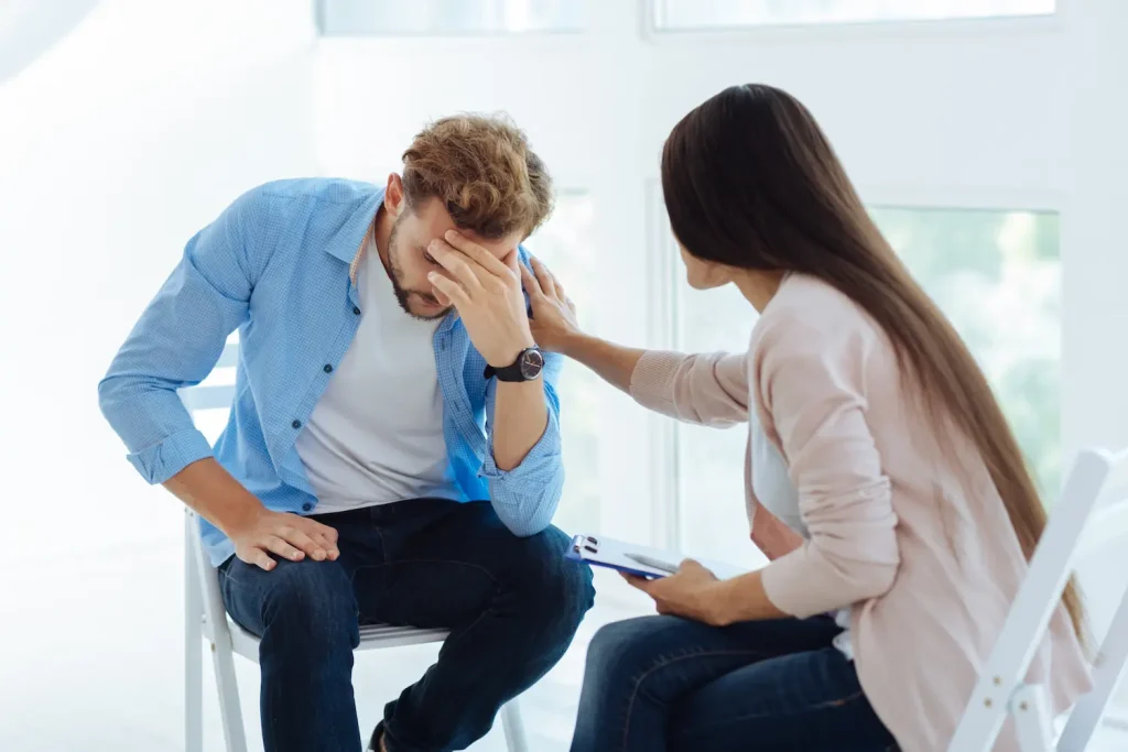 A woman holding a clipboard gently places a comforting hand on the shoulder of a distressed man sitting opposite her, who is looking down with his hand on his forehead.