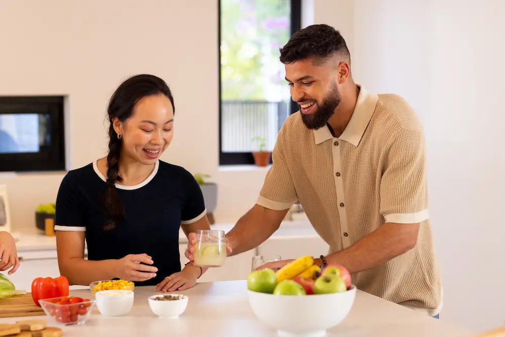 A smiling man and woman standing together at a bright kitchen island preparing a meal. The man is handing a glass of a light-colored drink to the woman across a counter filled with fresh fruit and bowls of chopped vegetables.