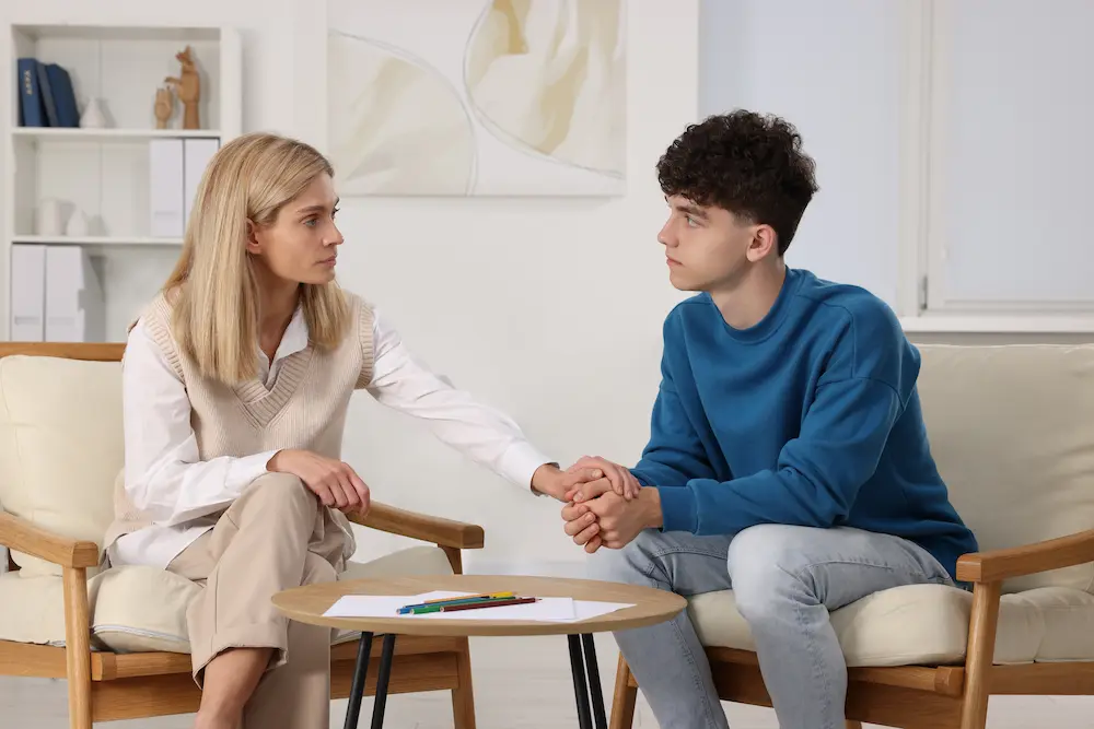 A blonde woman and a young man with curly dark hair sit facing each other in comfortable armchairs in a bright, modern office or counseling setting. The woman reaches across the space between them to gently hold the young man's hands in a supportive gesture. Both have serious expressions and are making direct eye contact. Between them is a small round table with a piece of paper and some colored pencils on it.